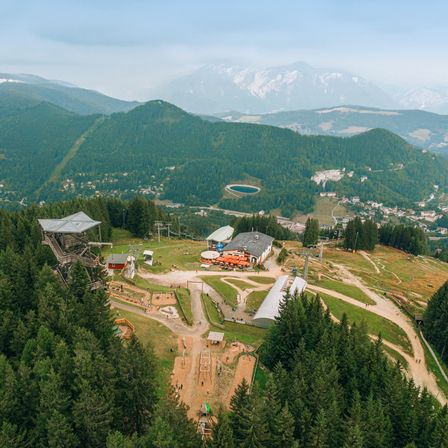 Luftaufnahme eines Skigebiets mit klarem blauem Himmel, schneebedeckten Bergen und einem dichten Wald. Verschiedene Strukturen, einschließlich eines Turms, Häuser und eine Straße, sind sichtbar.