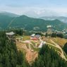 Aerial view of a ski resort with a clear blue sky, snow-capped mountains, and a dense forest. Various structures, including a tower, houses, and a road, are visible.