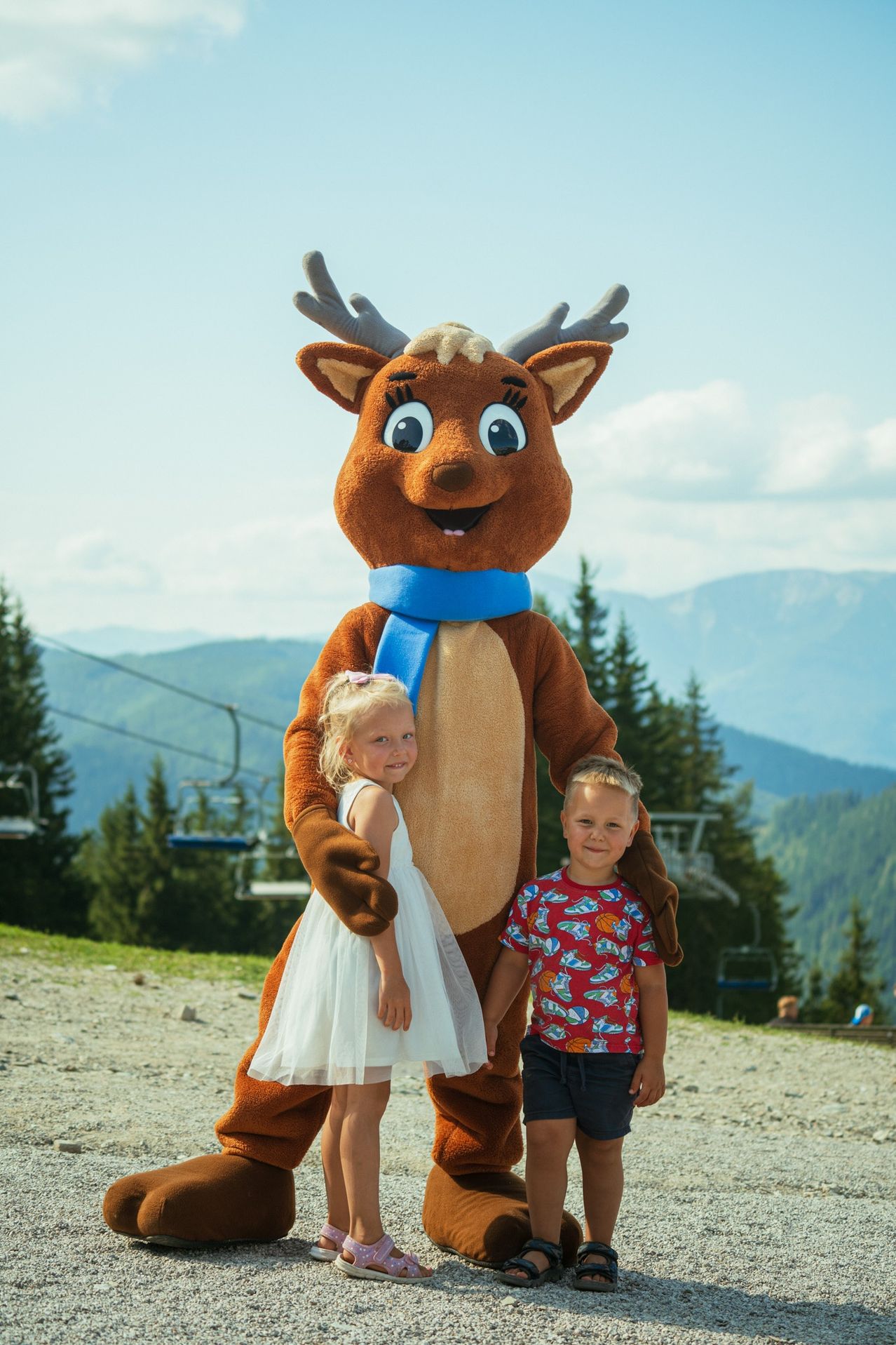 Ein Rentier-Maskottchen mit blauem Schal posiert mit zwei Kindern auf einem Bergweg, ein Skilift ist im Hintergrund zu sehen.