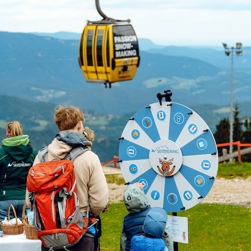 Eine Familie steht an einer Bergstation und schaut auf ein großes Drehrad. Sie sind in der Nähe einer Gondelbahn. In der Ferne sind Berge und ein klarer Himmel zu sehen.
