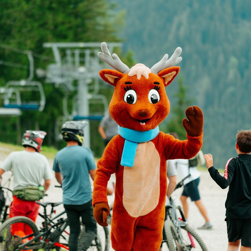 Ein Maskottchen, das als Rentier verkleidet ist, winkt einer Gruppe von Radfahrern in einer Berglandschaft zu.