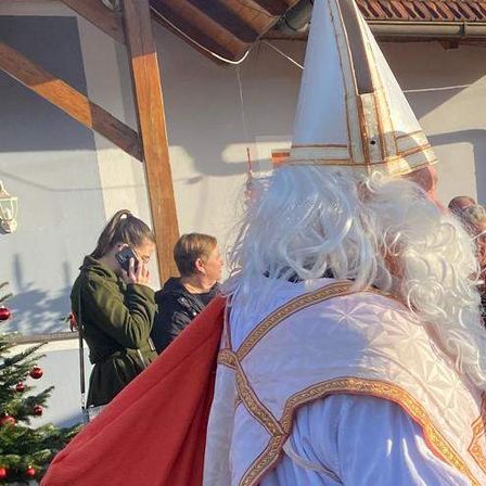 Ein Mann, der als Bischof gekleidet ist, spricht mit einer Frau vor einem Haus mit einem Weihnachtsbaum und mehreren Personen im Hintergrund.