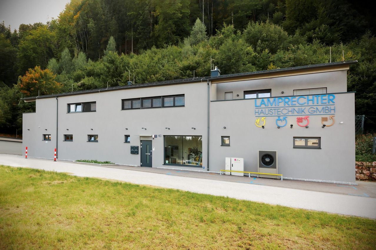 Ein graues Gebäude namens Lamprecht Haustechnik vor einem Wald mit vielen Fenstern und einer Glastür.