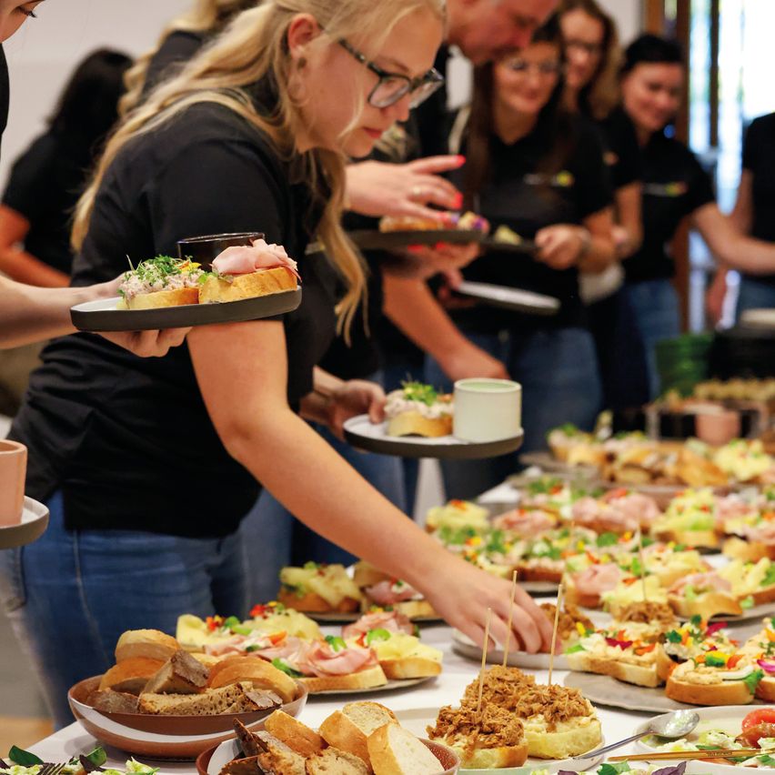 Eine Gruppe von Menschen in schwarzen Shirts serviert Essen auf Tellern. Das Essen umfasst Sandwiches und andere Gerichte. Der Tisch ist voll mit verschiedenen Arten von Essen.