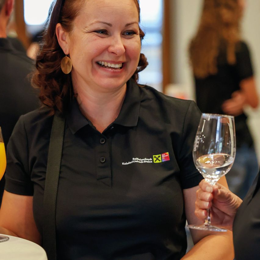 Eine lächelnde Frau in einem schwarzen Shirt hält ein Weinglas und steht bei einer Veranstaltung. Eine andere Person hält ein Glas Orangensaft.