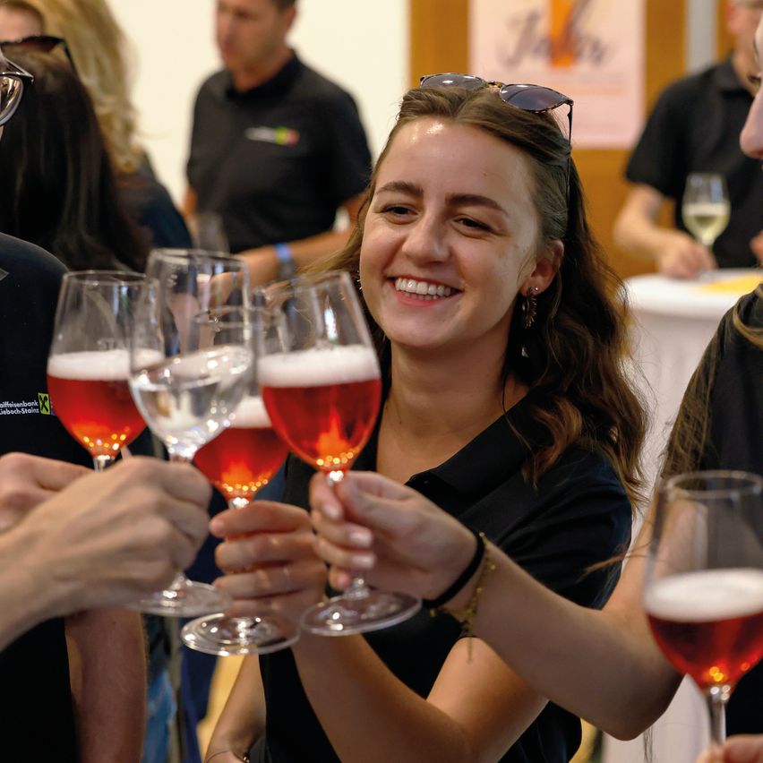 Eine Gruppe von Menschen hält Gläser mit Bier in die Höhe und lächelt. Eine Frau mit einer Brille auf dem Kopf schaut direkt in die Kamera.