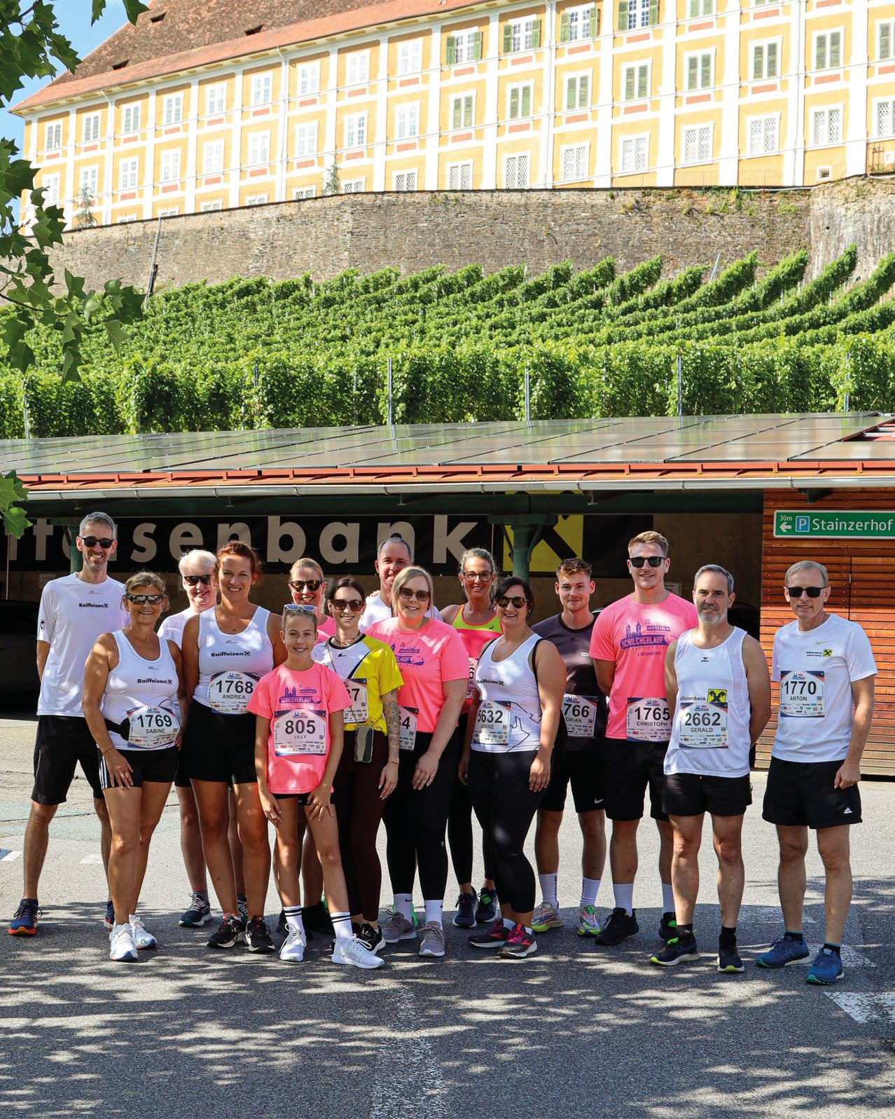 Eine Gruppe von Läufern in identischen weißen und rosa Shirts steht vor einem Gebäude mit Solarpanelen und einem Weinberg im Hintergrund.