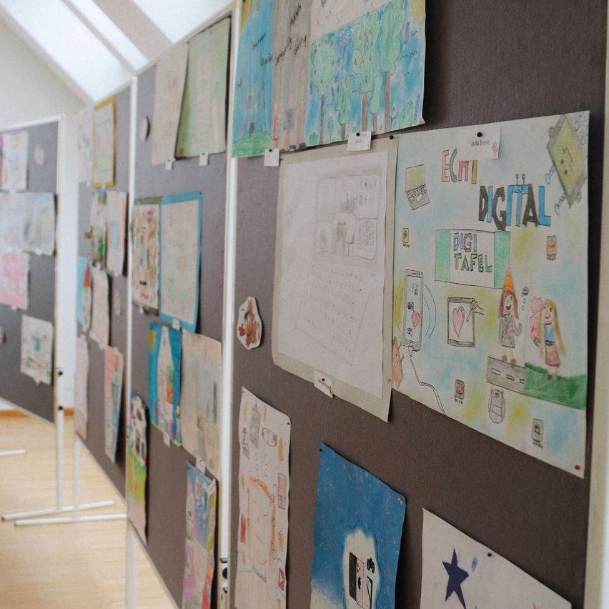 Eine Wand mit vielen bunten Kinderzeichnungen, einige mit Etiketten befestigt, auf einem Holzboden. Die Zeichnungen umfassen Themen wie Technologie und Natur.