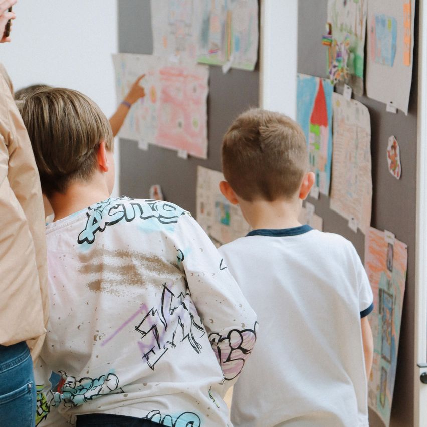 Zwei Jungen betrachten Kunstwerke an einer Wand, einer mit einem Graffiti-Stil-Shirt, während ein anderer auf ein Werk zeigt. Eine Person in Jeans steht neben ihnen.