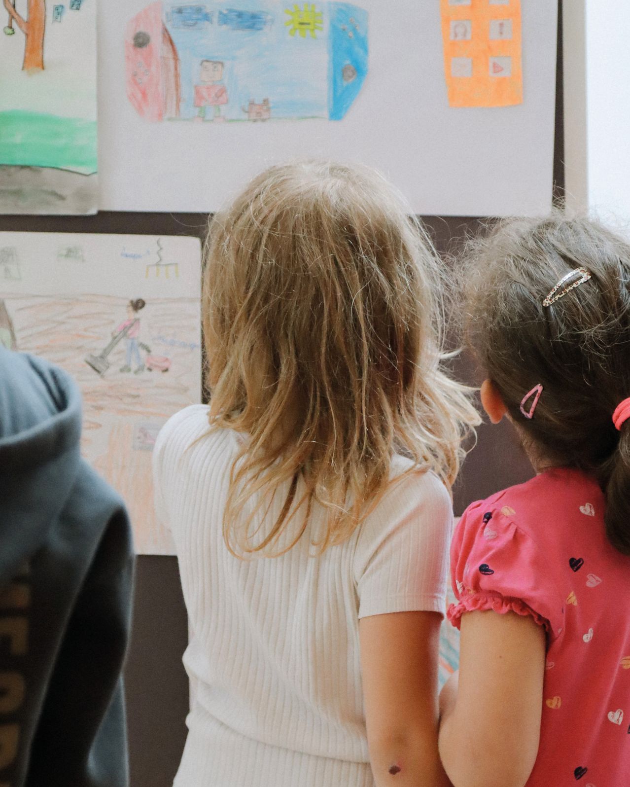 Drei Kinder stehen neben einer mit Zeichnungen bedeckten Wand. Das Mädchen auf der rechten Seite trägt ein rosa Shirt mit Herz-Designs. Das Mädchen in der Mitte trägt ein weißes Shirt.