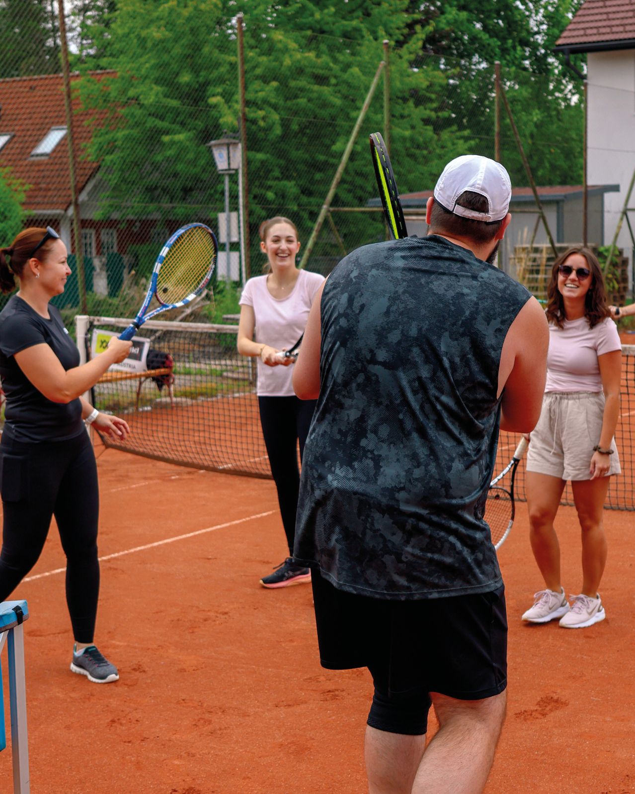 Drei Personen sind auf einem Tennisplatz. Ein Mann und zwei Frauen halten Tennisschläger, und der Mann schaut die Frauen an.