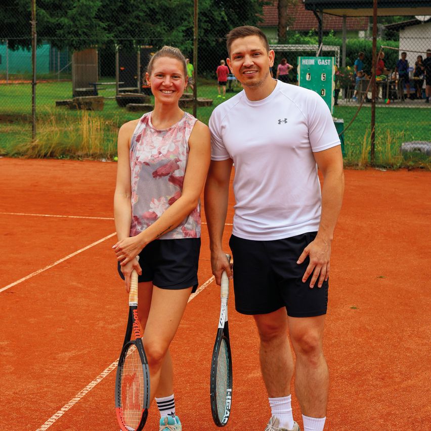 Ein Mann und eine Frau stehen auf einem Tennisplatz, lächeln und halten Tennisschläger in der Hand. Hinter ihnen umgibt ein Zaun den Platz, und ein Spielfeld ist sichtbar. Im Hintergrund ist ein Rasenbereich mit Menschen und Bäumen zu sehen.