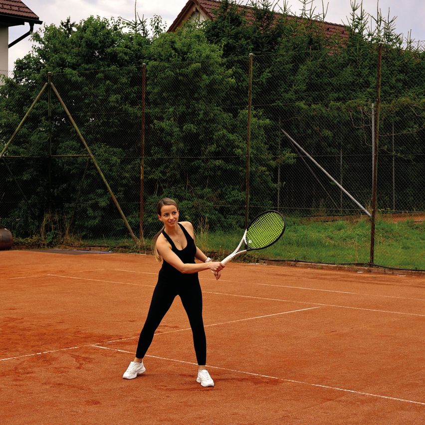 Eine Frau in schwarzer Tenniskleidung schwingt einen Schläger auf einem Tennisplatz, mit einem Zaun und Bäumen im Hintergrund.