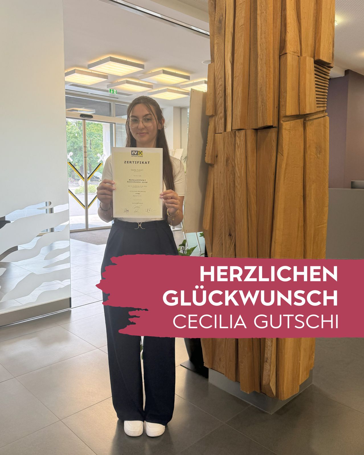 Eine Frau steht in einem Büro und hält ein Zertifikat in der Hand. Hinter ihr steht eine große Holzskulptur. Über ihr steht 'HERZLICHEN GLUCKWUNSCH CECILIA GUTSCHI'.