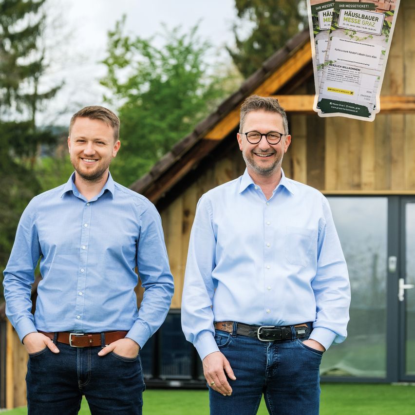 Zwei Männer in blauen Hemden und Jeans stehen vor einem Haus, lächeln und posieren für ein Foto. Hinter ihnen ist ein Plakat mit Text in Deutsch.