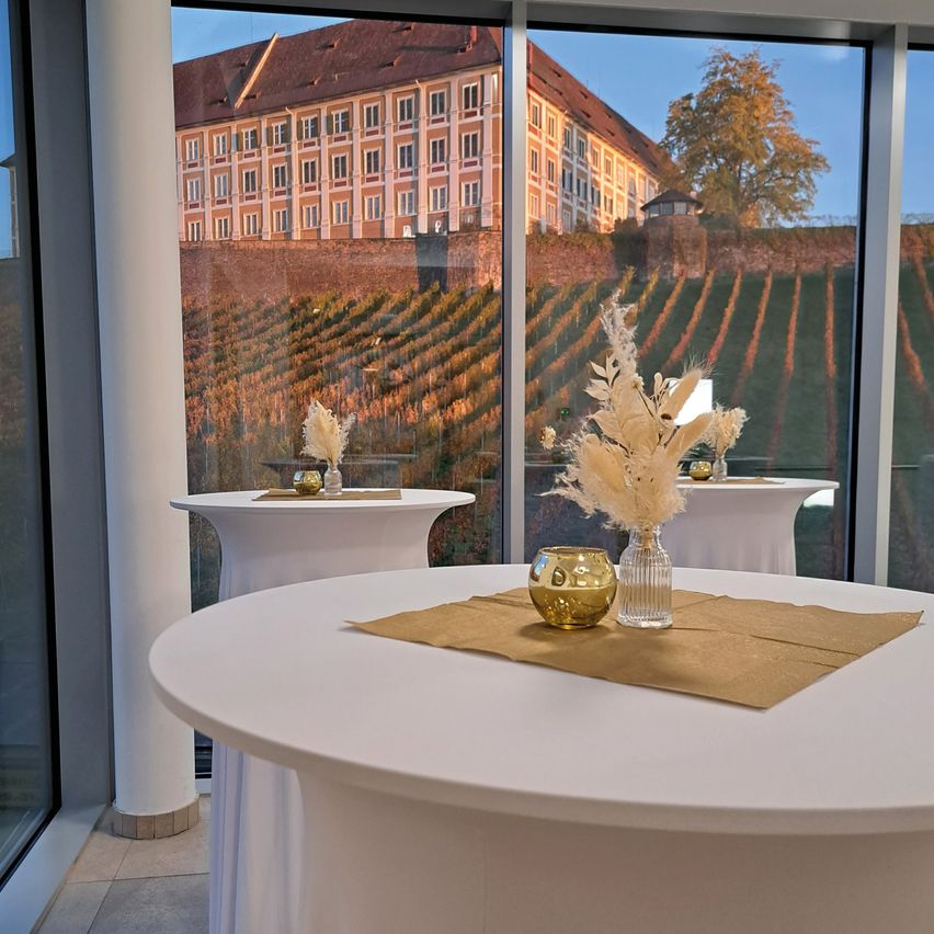 Eine Inneneinrichtung mit runden Tischen und Glaswänden, die ein Weingut und ein Gebäude im Hintergrund reflektiert.