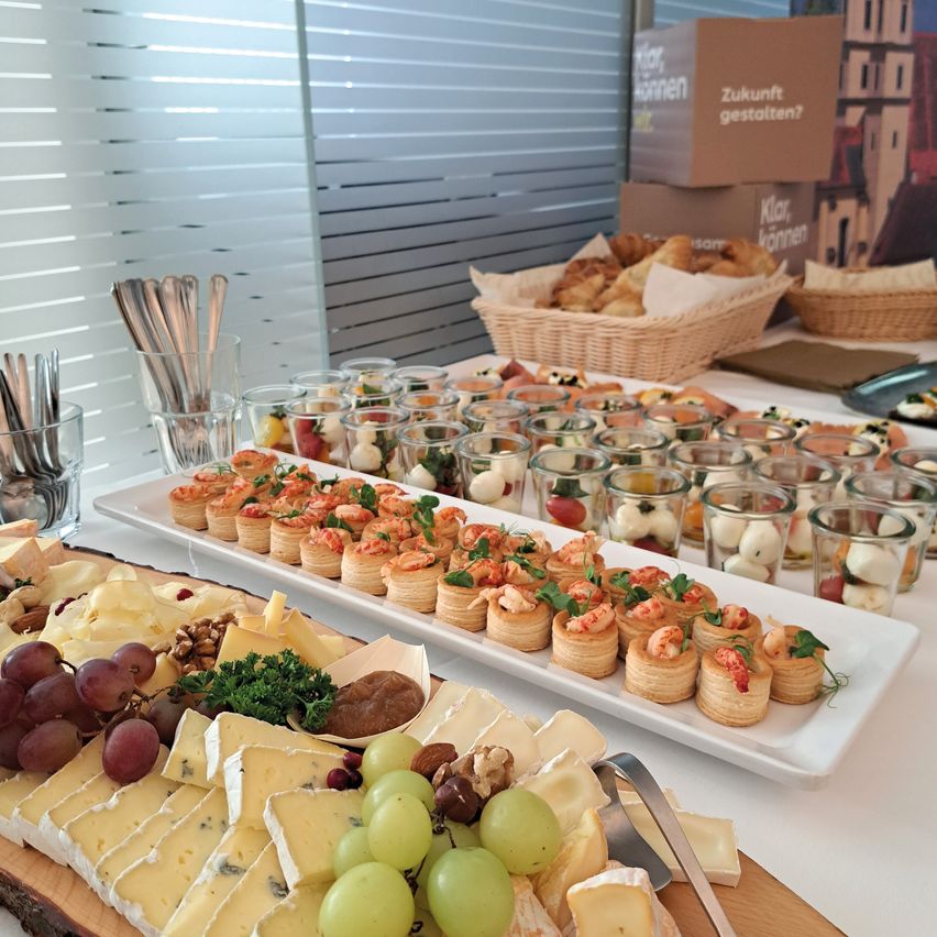 Ein weißer Tisch zeigt eine Vielzahl von Lebensmitteln. Auf Holzbrettern sind Teller mit Käse, Trauben und Nüssen angeordnet. Es gibt auch kleine Törtchen und verschiedene Vorspeisen in Gläsern. Im Hintergrund sind Kisten und ein Korb mit Brot zu sehen.