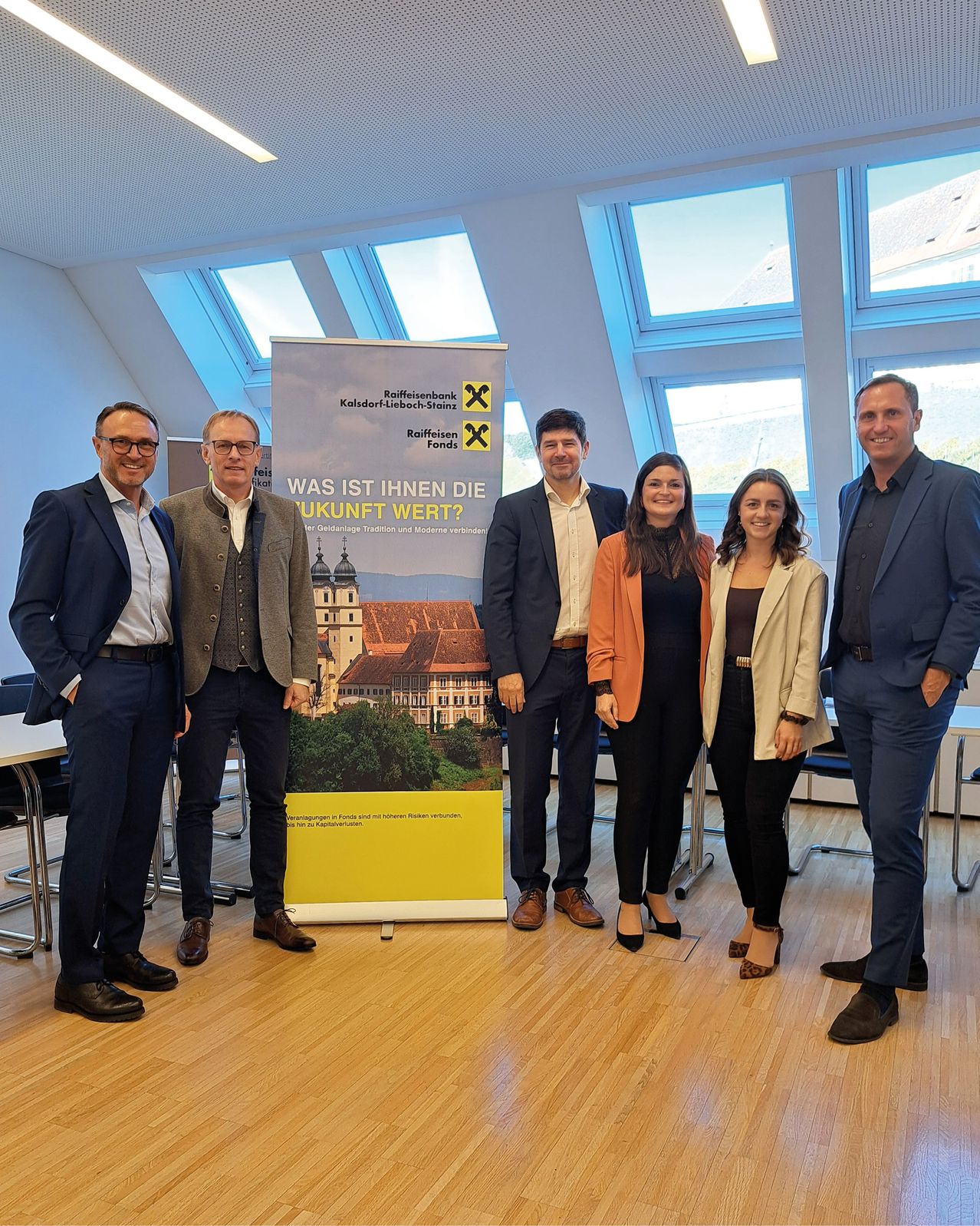 Fünf Personen stehen vor einem Banner mit dem Text 'Was ist Ihnen die Zukunft wert?' Eine Frau mit einer Kamera macht ein Foto von ihnen.