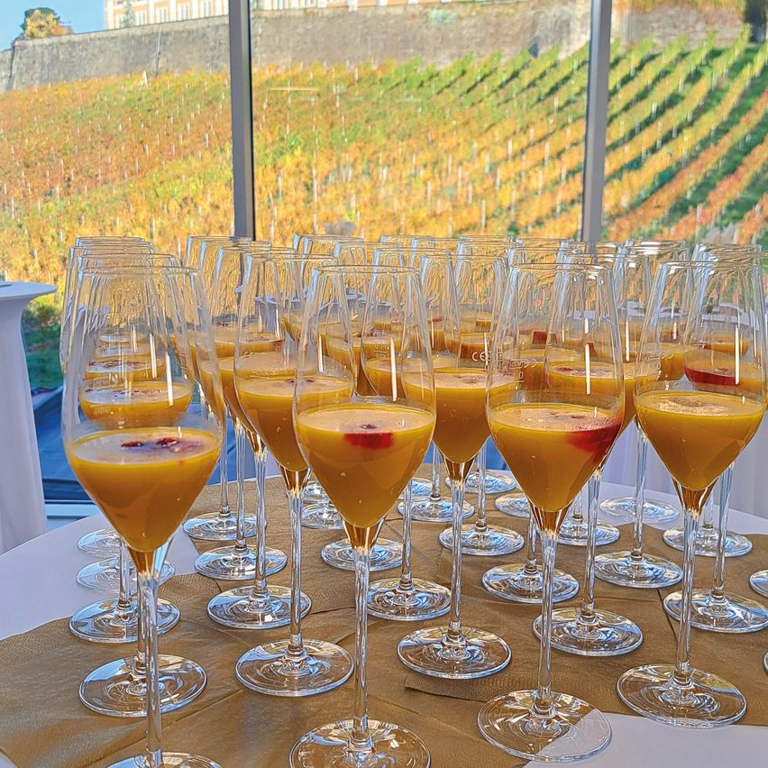 Mehrere Gläser mit orangefarbenen Getränken sind auf einem Tisch angeordnet, mit Blick auf ein Weingut im Hintergrund.