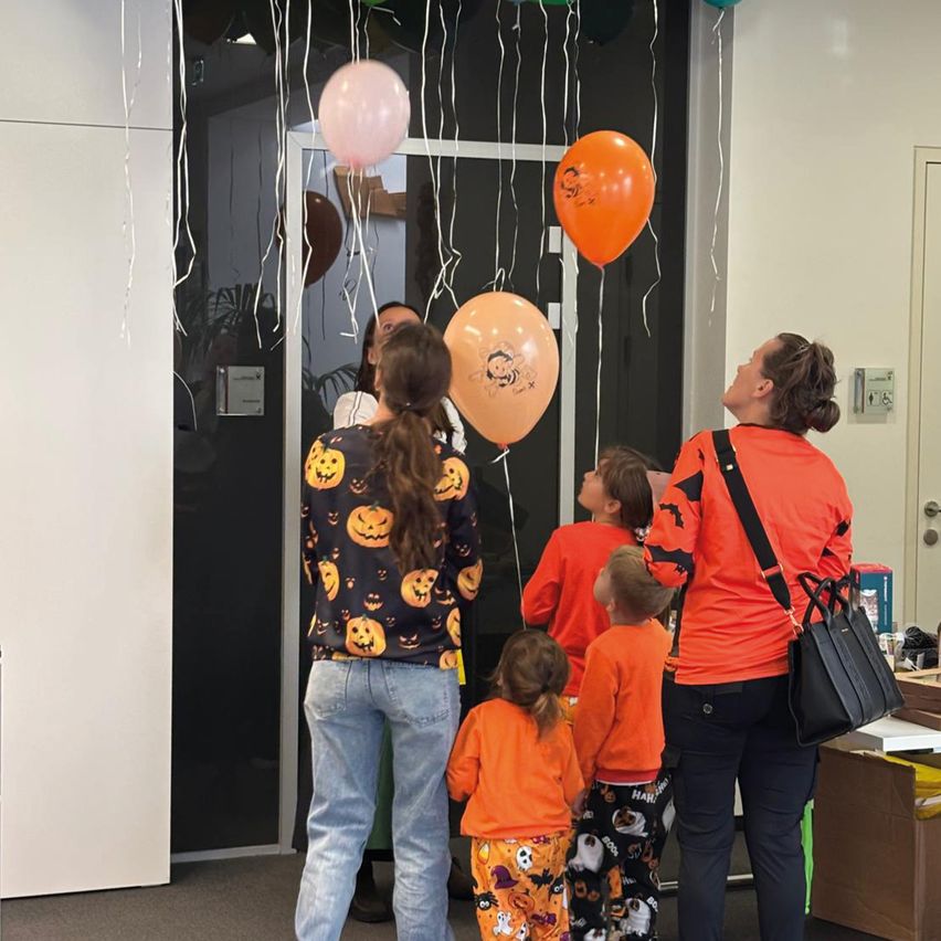 Eine Gruppe von Menschen, einschließlich Erwachsener und Kindern, ist drinnen. Einige schauen zu Luftballons auf, die von der Decke hängen. Die Kinder tragen orangefarbene Hemden und die Erwachsenen orangefarbene und schwarze Hemden.
