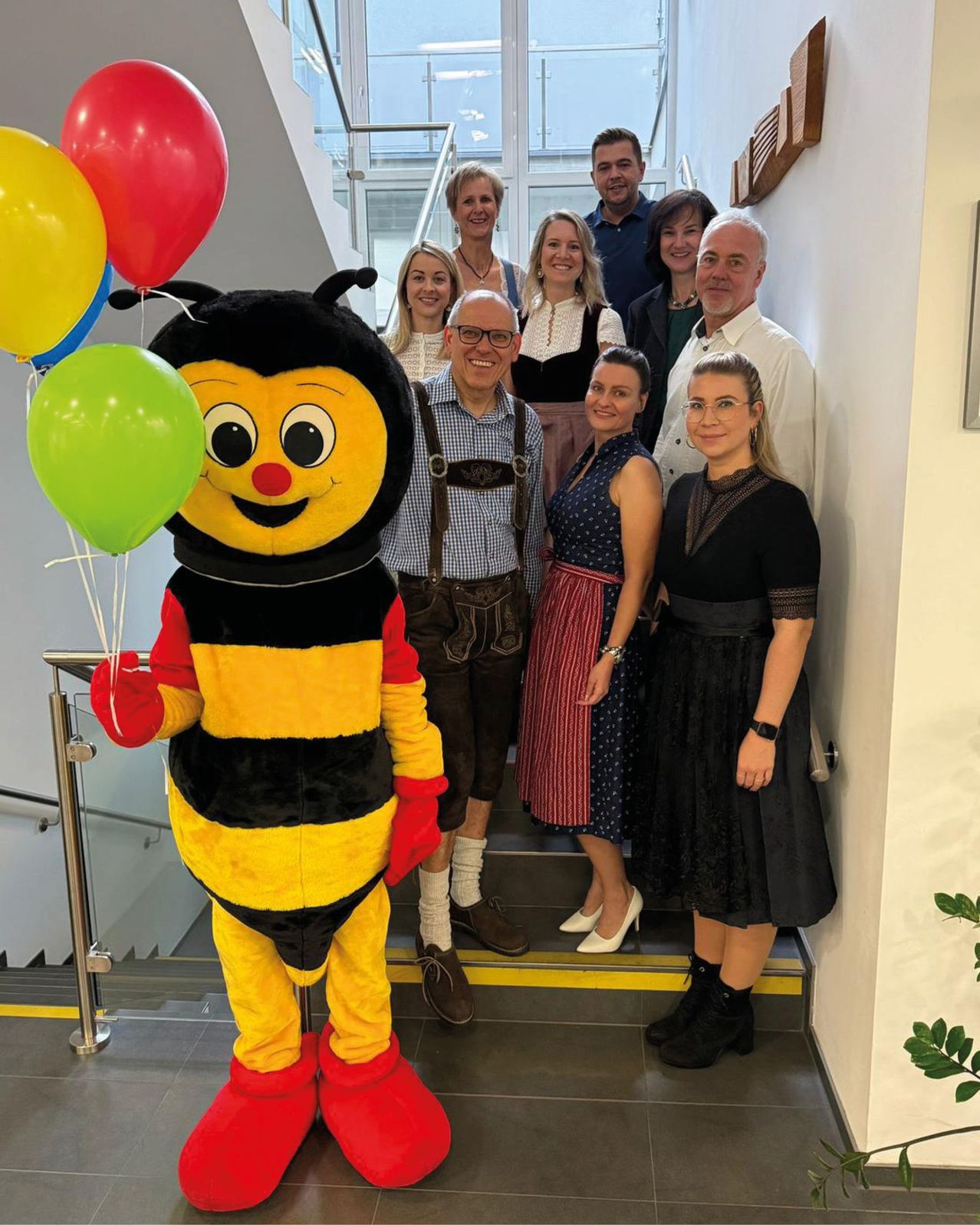 Eine Gruppe von Menschen in traditioneller Kleidung posiert für ein Foto auf einer Treppe mit einem Bienen-Maskottchen, das Luftballons hält.