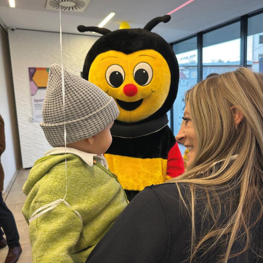 Eine lächelnde Frau hält ein kleines Kind, das einen großen Bienen-Mascot mit roter Nase ansieht. Sie sind drinnen in der Nähe einer Glaswand.