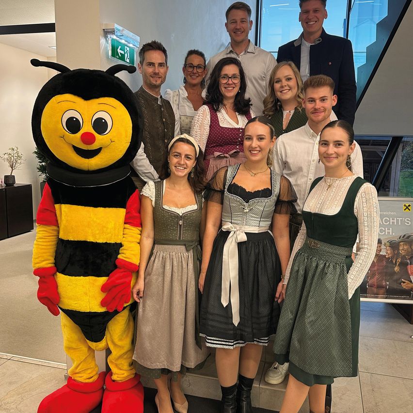 Eine Gruppe von Menschen in traditioneller bayerischer Kleidung, einschließlich Dirndln, posiert für ein Foto mit einem Biene-Maskottchen in einer Lobby. Dahinter sind ein großes Fenster und ein Schild mit einem Ausgangssymbol sichtbar.