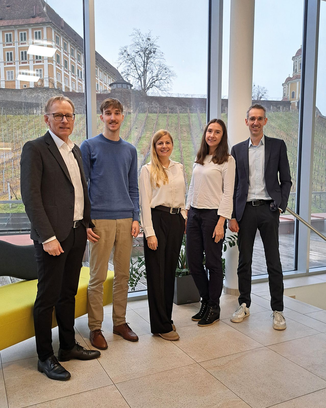 Fünf Personen, drei Männer und zwei Frauen, posieren für ein Foto in einem Innenraum mit großen Glasfenstern. Sie tragen formelle Kleidung.