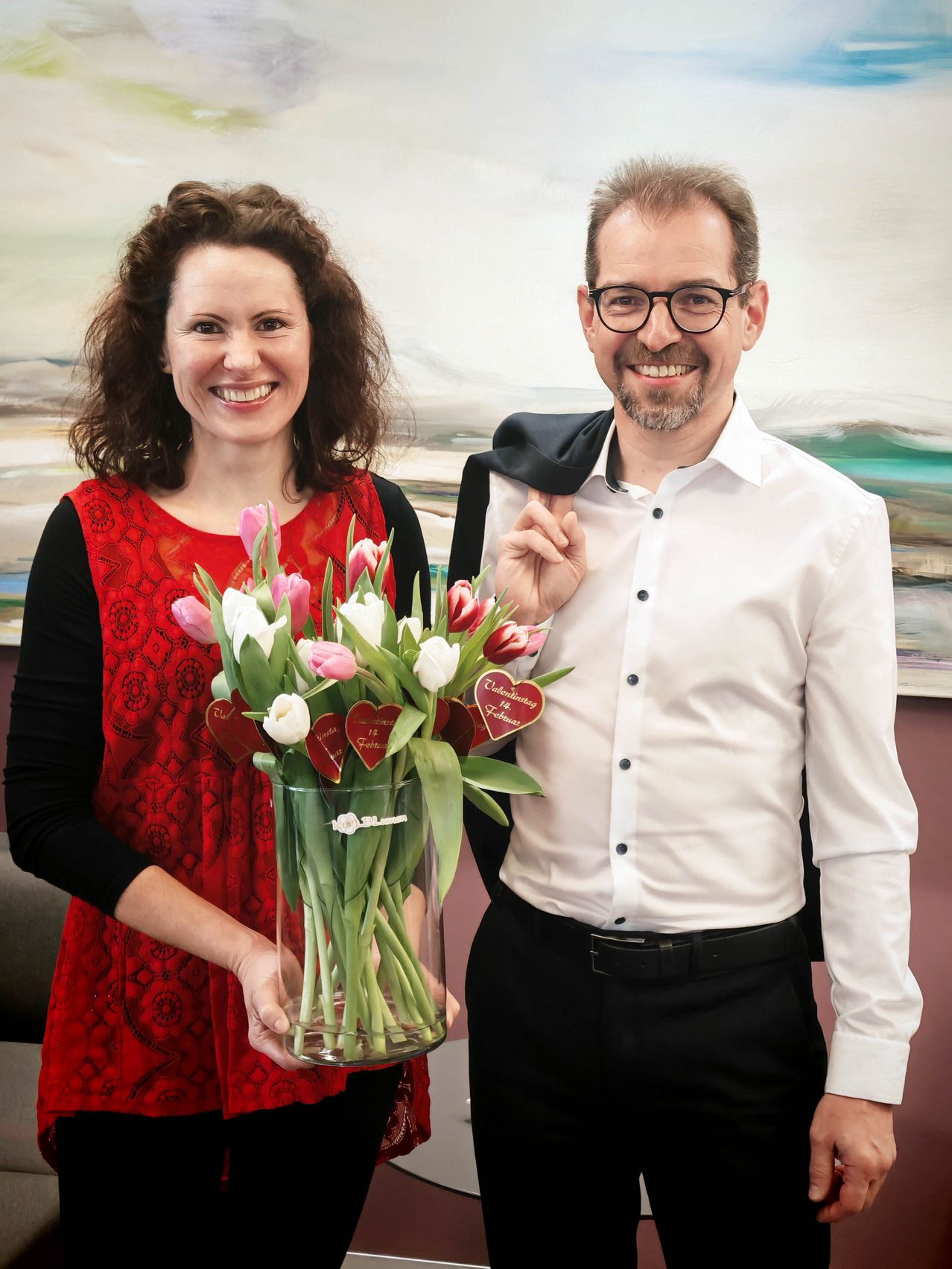A man and a woman stand next to each other, smiling. The woman holds a vase with pink and white tulips. They both have heart-shaped tags on their clothes. The man is wearing a white shirt and glasses.