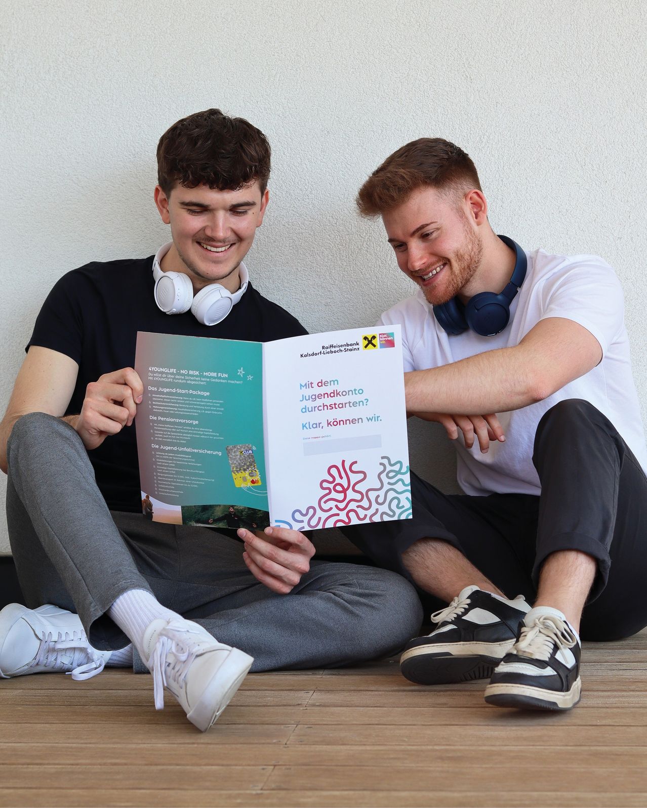 Zwei junge Männer sitzen auf dem Boden, einer liest eine Broschüre mit einem lächelnden Gesicht. Die Broschüre hat ein farbenfrohes Design mit Text und Illustrationen. Der Mann rechts trägt Kopfhörer.