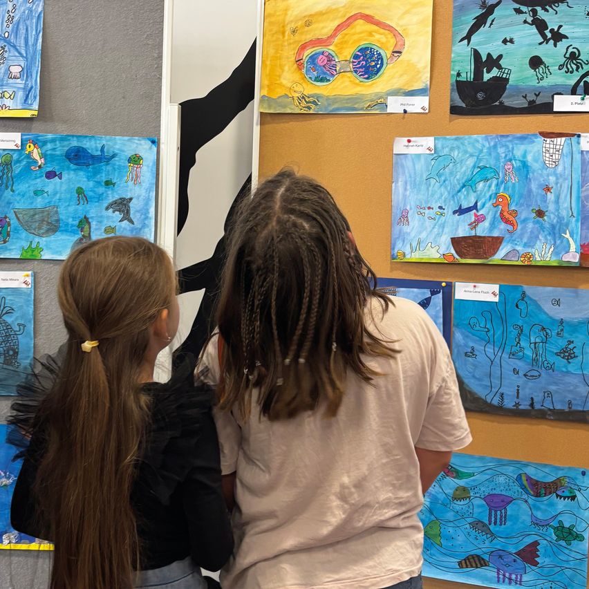 Two girls stand in front of a wall with various paintings, one of which is of a whale. They look at the paintings, with one girl having long hair.
