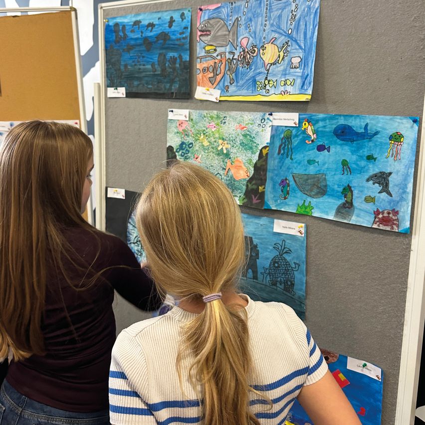 Two girls with long hair view marine life paintings on a gray wall. Various art pieces depict fish, sea creatures, and underwater scenes.