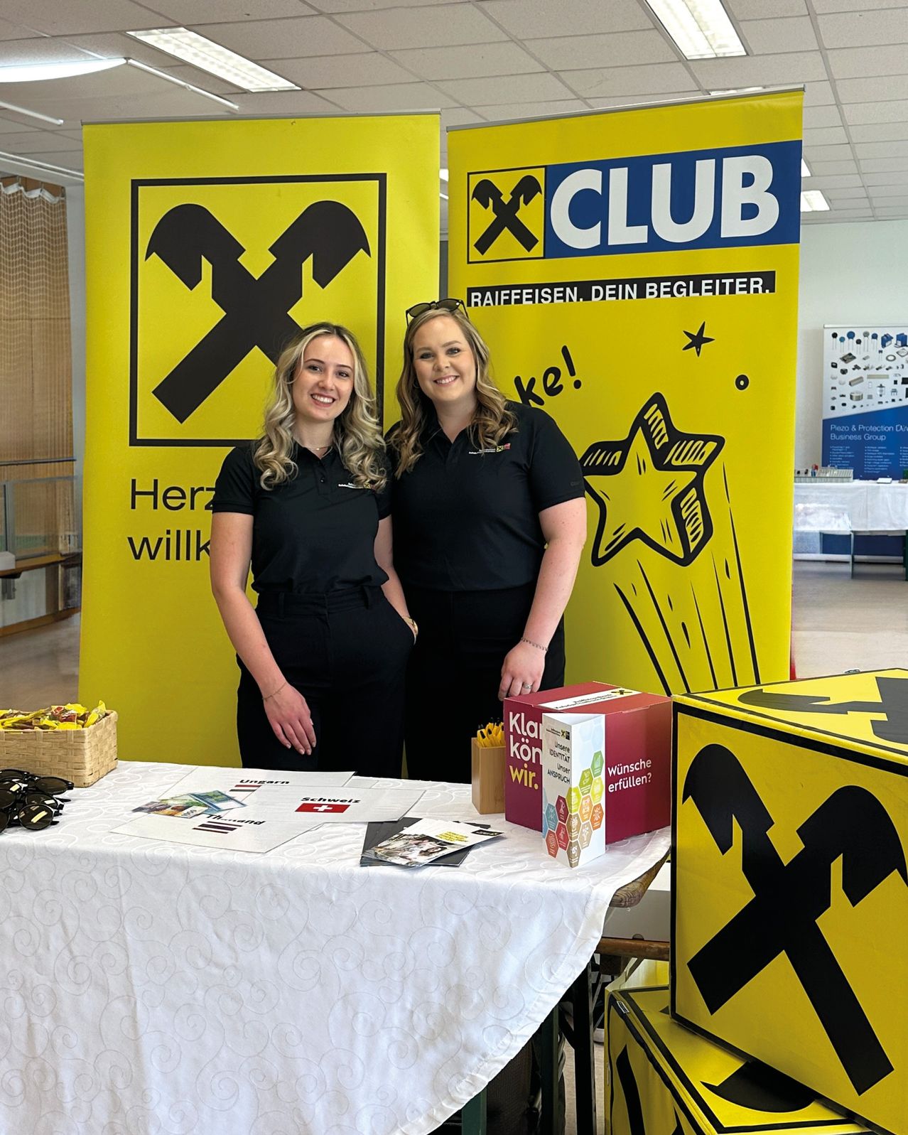 Two women stand behind a table with promotional items. Behind them, large yellow banners with the X-Club logo and text are displayed.