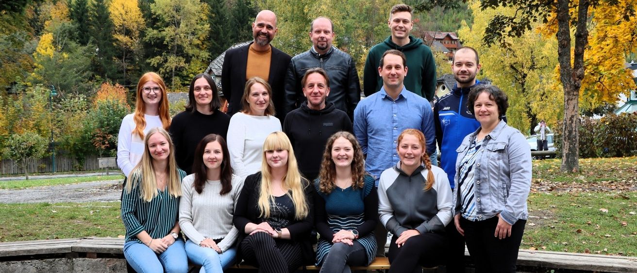 Eine Gruppe von Menschen, sowohl Männer als auch Frauen, lächeln und posieren für ein Foto auf einer Bank im Freien. Sie befinden sich in einem Park mit Bäumen.