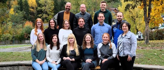 Eine Gruppe von Menschen, sowohl Männer als auch Frauen, lächeln und posieren für ein Foto auf einer Bank im Freien. Sie befinden sich in einem Park mit Bäumen.