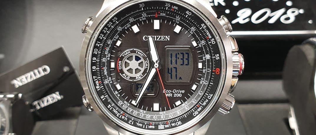 Eine silberne Citizen Eco-Drive Uhr wird auf schwarzem Hintergrund präsentiert. Die Uhr hat ein schwarzes Zifferblatt mit weißen und roten Akzenten. Die Zeit ist 11:31 Uhr.