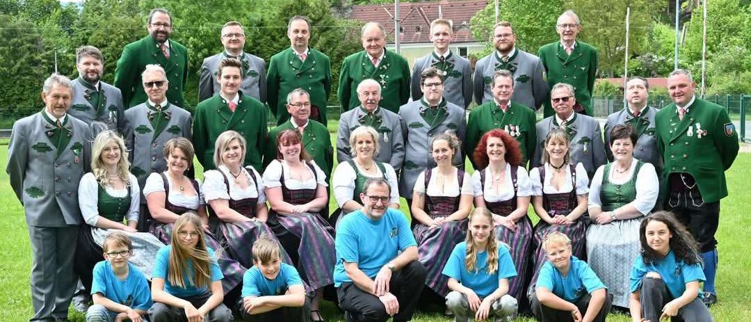 Eine Gruppe von Menschen in traditioneller bayerischer Kleidung, einschließlich grüner und weißer Jacken, posiert für ein Foto auf einem Grasfeld. Dahinter stehen Bäume und ein Haus.