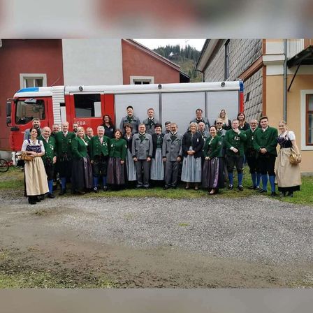 Eine Gruppe von Menschen in grünen und weißen Trachten steht vor einem roten Feuerwehrauto und posiert für ein Foto. Im Hintergrund befinden sich Gebäude.