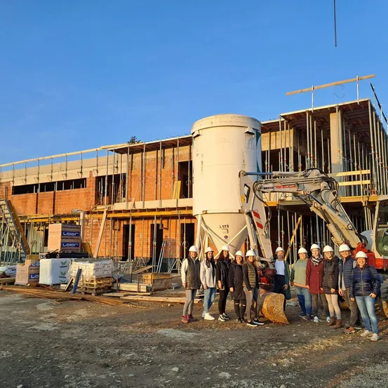 Eine Gruppe von Menschen in Schutzhelmen steht vor einer Baustelle mit einem großen im Bau befindlichen Gebäude und einem nahegelegenen Baufahrzeug.