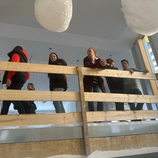 Eine Gruppe von Menschen steht auf einem hölzernen Balkon und schaut nach unten auf den Boden. Sie tragen Winterkleidung. Die Decke hat weiße Kunststofflampen.