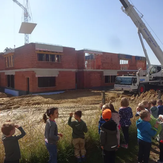 Kinder beobachten eine Baustelle mit einem Kran, der einen großen Ziegel hebt. Das Gebäude wird gebaut, und die Kinder stehen auf einem Feld.
