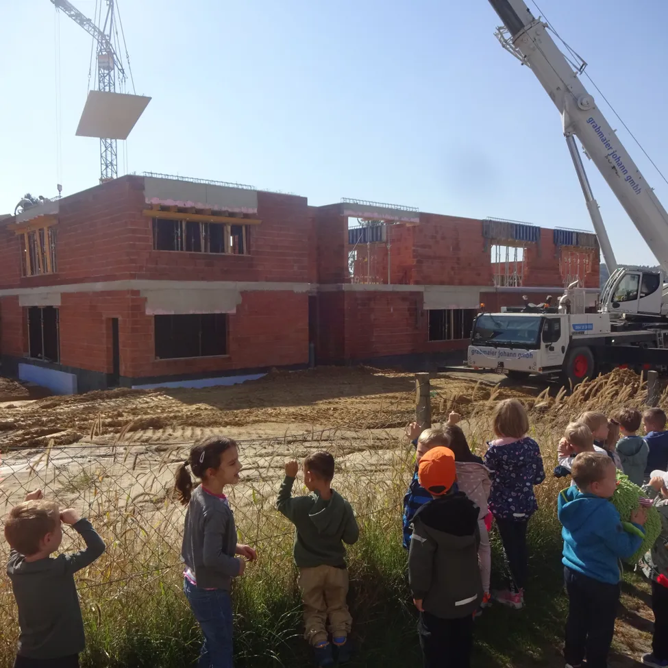 Kinder beobachten eine Baustelle mit einem Kran, der einen großen Ziegel hebt. Das Gebäude wird gebaut, und die Kinder stehen auf einem Feld.