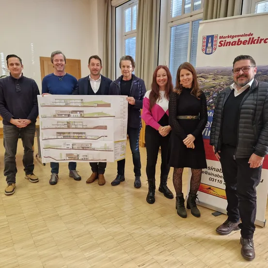 Eine Gruppe von Menschen steht in einem Raum und hält einen großen Architekturplan. Sie lächeln und scheinen für ein Foto zu posieren. Hinter ihnen ist ein Banner mit einem Logo und Text zu sehen.