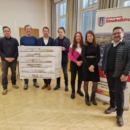Eine Gruppe von Menschen steht in einem Raum und hält einen großen Architekturplan. Sie lächeln und scheinen für ein Foto zu posieren. Hinter ihnen ist ein Banner mit einem Logo und Text zu sehen.