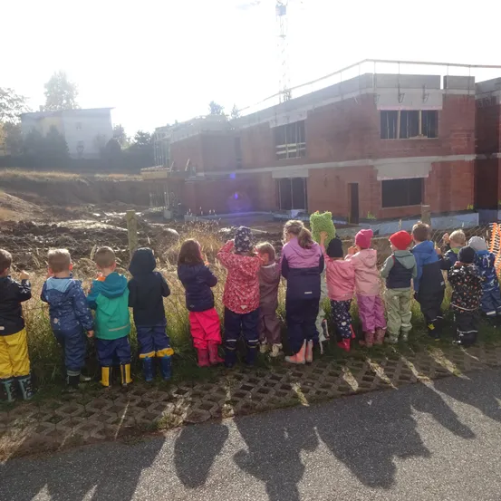 Eine Gruppe von Kindern in Regenmänteln und Stiefeln steht in einer Reihe und schaut auf eine Baustelle.