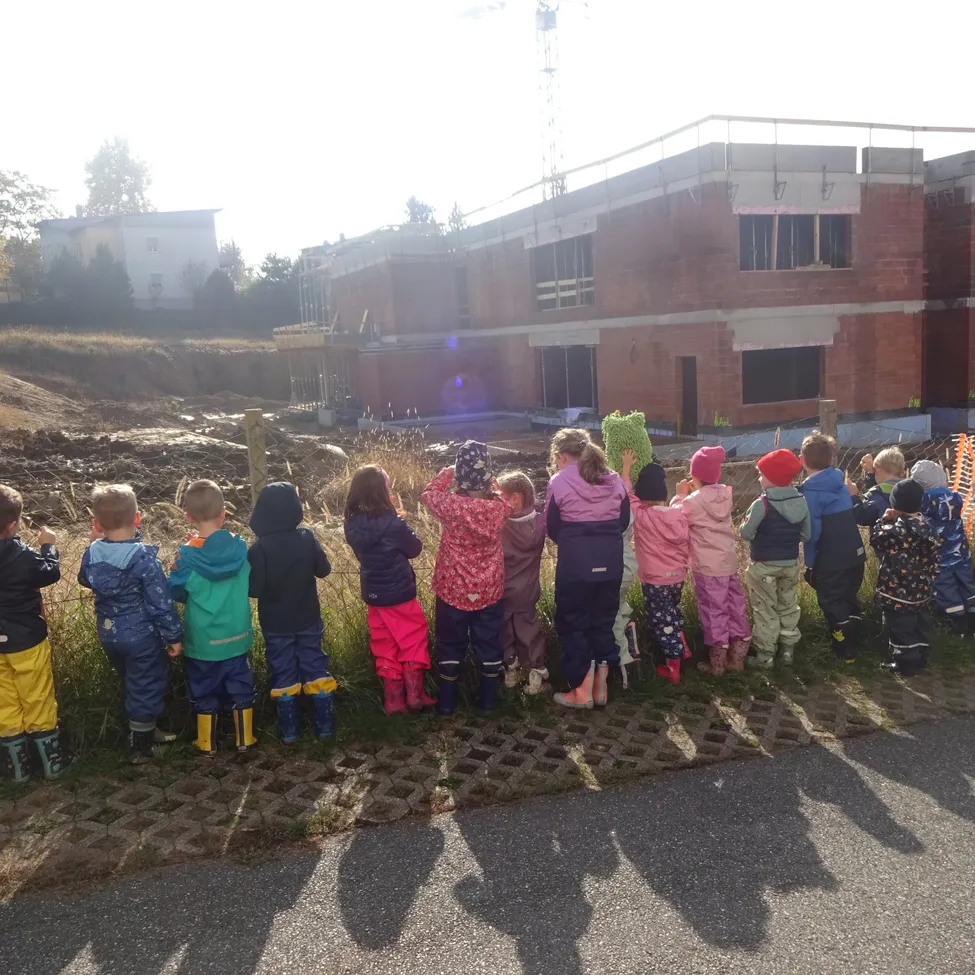 Eine Gruppe von Kindern in Regenmänteln und Stiefeln steht in einer Reihe und schaut auf eine Baustelle.