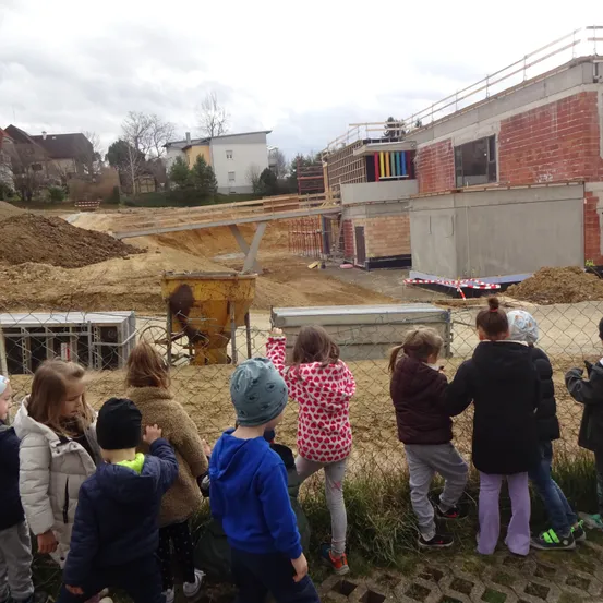 Eine Gruppe von Kindern beobachtet eine Baustelle. Ein gelber Baumaschinen steht in der Nähe. Die Kinder stehen hinter einem Zaun. Dahinter ist ein im Bau befindliches Gebäude zu sehen.