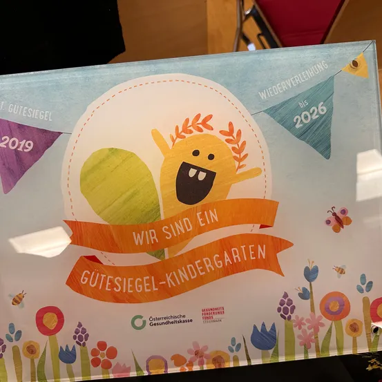 Ein farbenfrohes Poster mit einer Cartoon-Figur, Blumen und einem Band, auf dem 'Wir sind ein Gutesiegel-Kindergarten' steht. Das Poster enthält auch die Jahre 2019 und 2026.