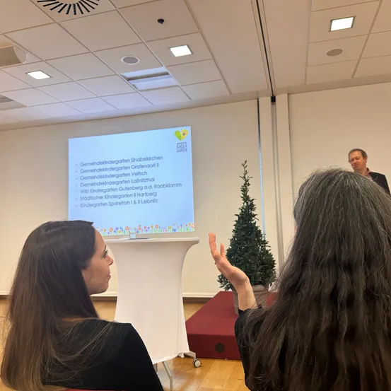 Zwei Frauen sitzen in einem Raum und sehen einen Mann, der vor einem Podium steht. Neben dem Podium steht ein Weihnachtsbaum und eine Projektionsleinwand zeigt eine Liste von Kindergärten.