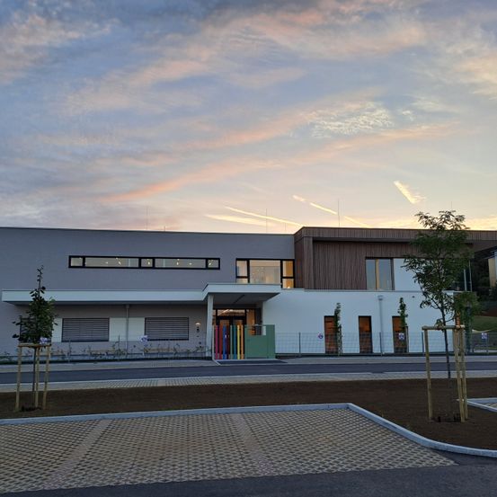 Ein modernes Gebäude mit einem farbenfrohen Eingang ist unter einem bewölkten Sonnenuntergang zu sehen. Bäume und Pflanzen sind im Vordergrund.